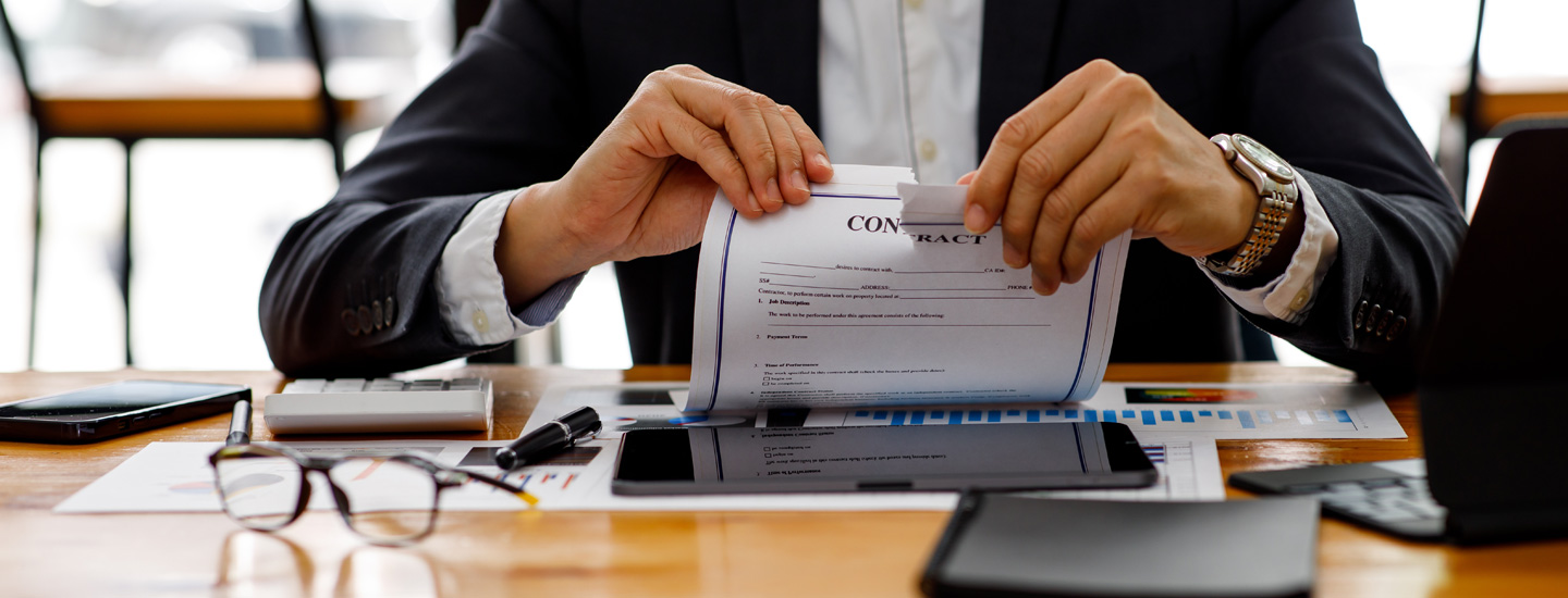 Businessperson tearing a contract document in an office setting, with a calculator, tablet, phone, and paperwork on the desk.