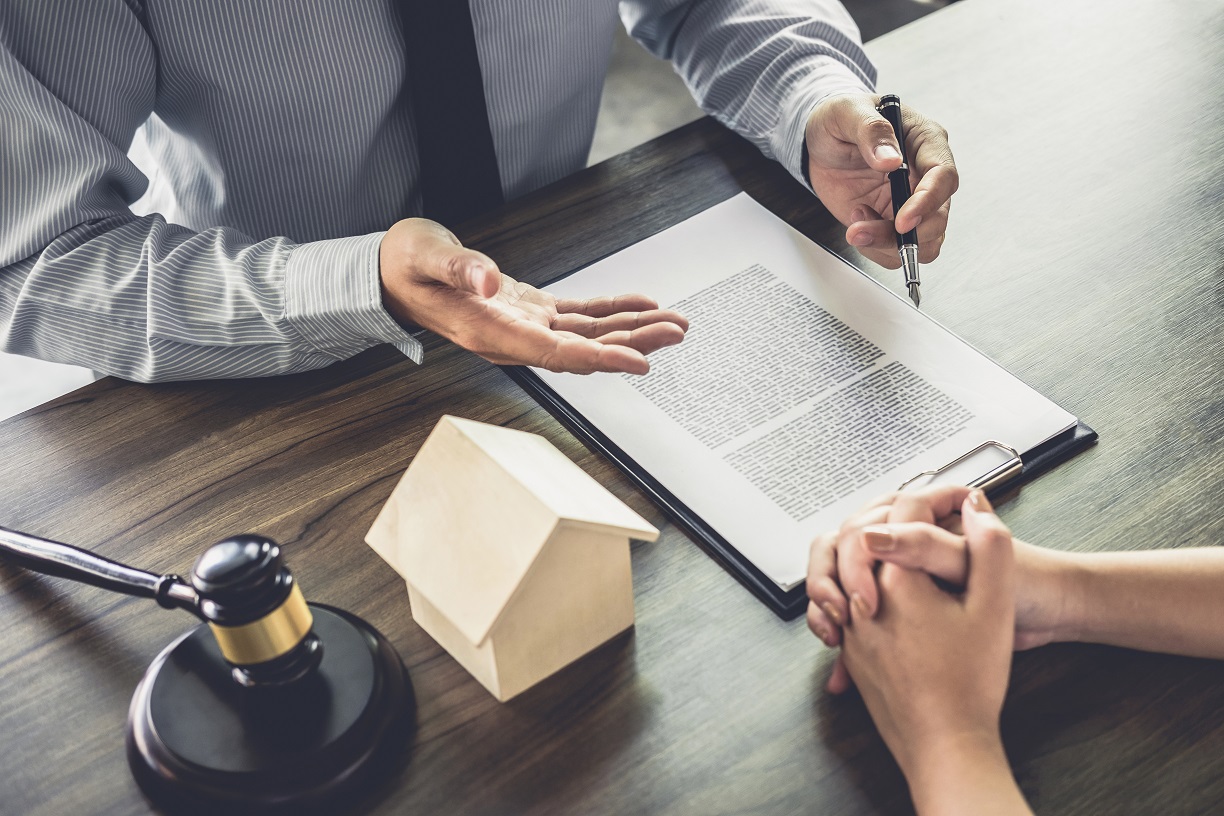 A real estate agent explaining a contract to a client at a desk, with a model house, gavel, and document folder on the table.
