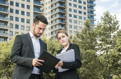 Professional businessman and businesswoman reviewing documents and holding a folder in front of a modern office building.