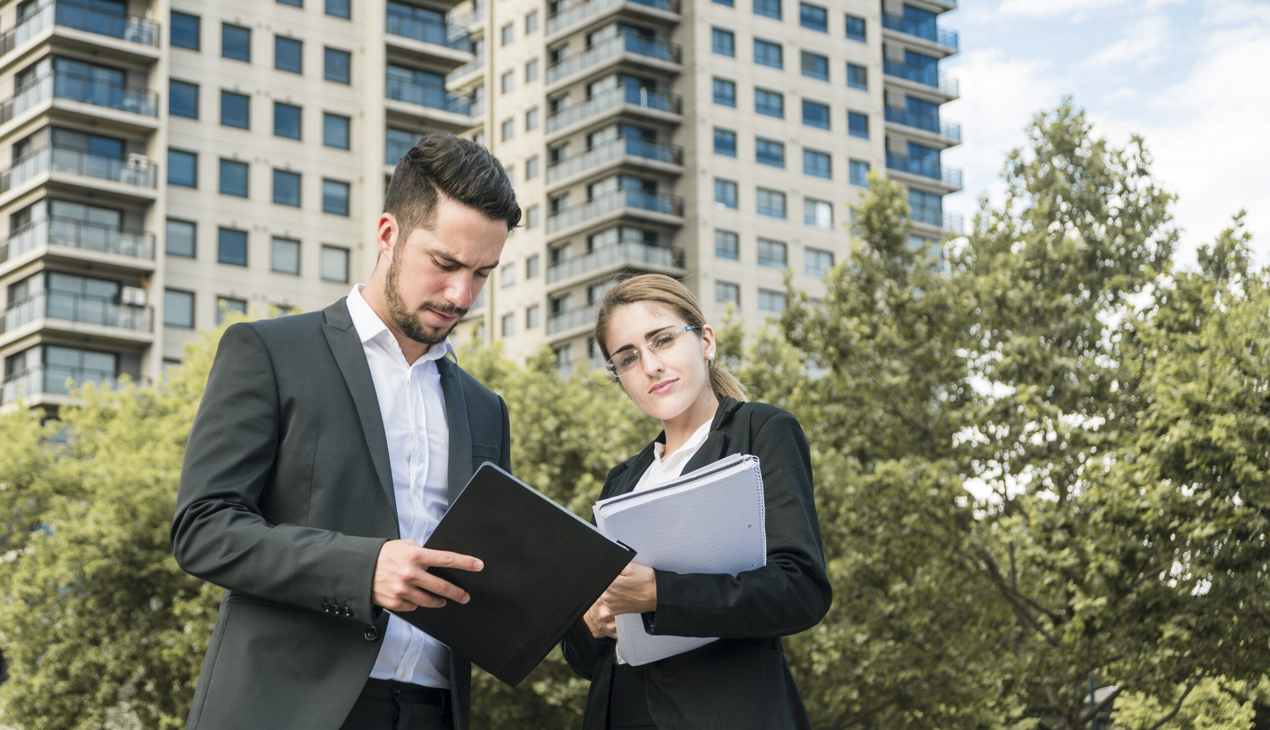 Professional businessman and businesswoman reviewing documents and holding a folder in front of a modern office building.
