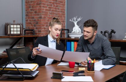 top-view-young-busy-motivated-clerical-workers-discussing-one-issue-documents-office Two young professional office workers actively discussing legal documents and project strategies at a wooden desk with office supplies and a brick wall background.