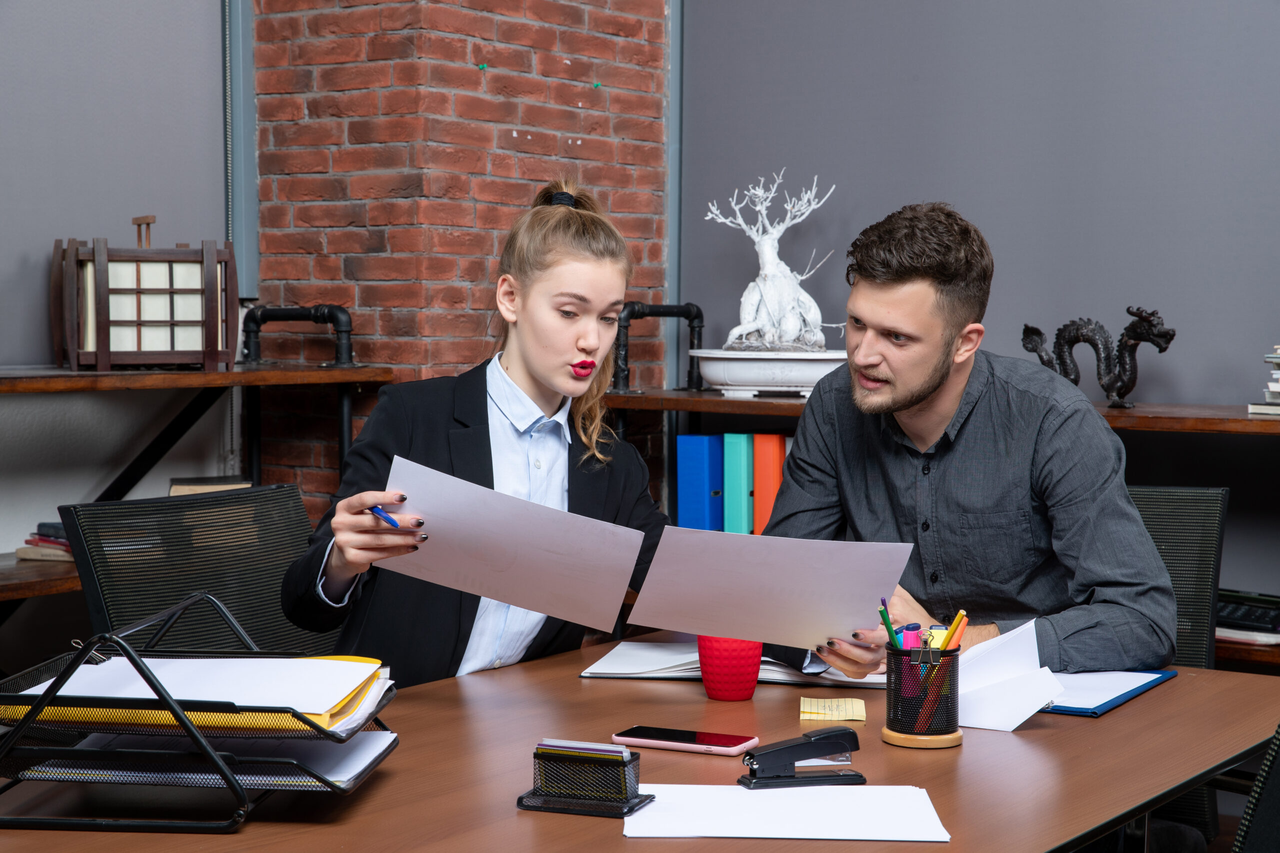 Two young professional office workers actively discussing legal documents and project strategies at a wooden desk with office supplies and a brick wall background.
