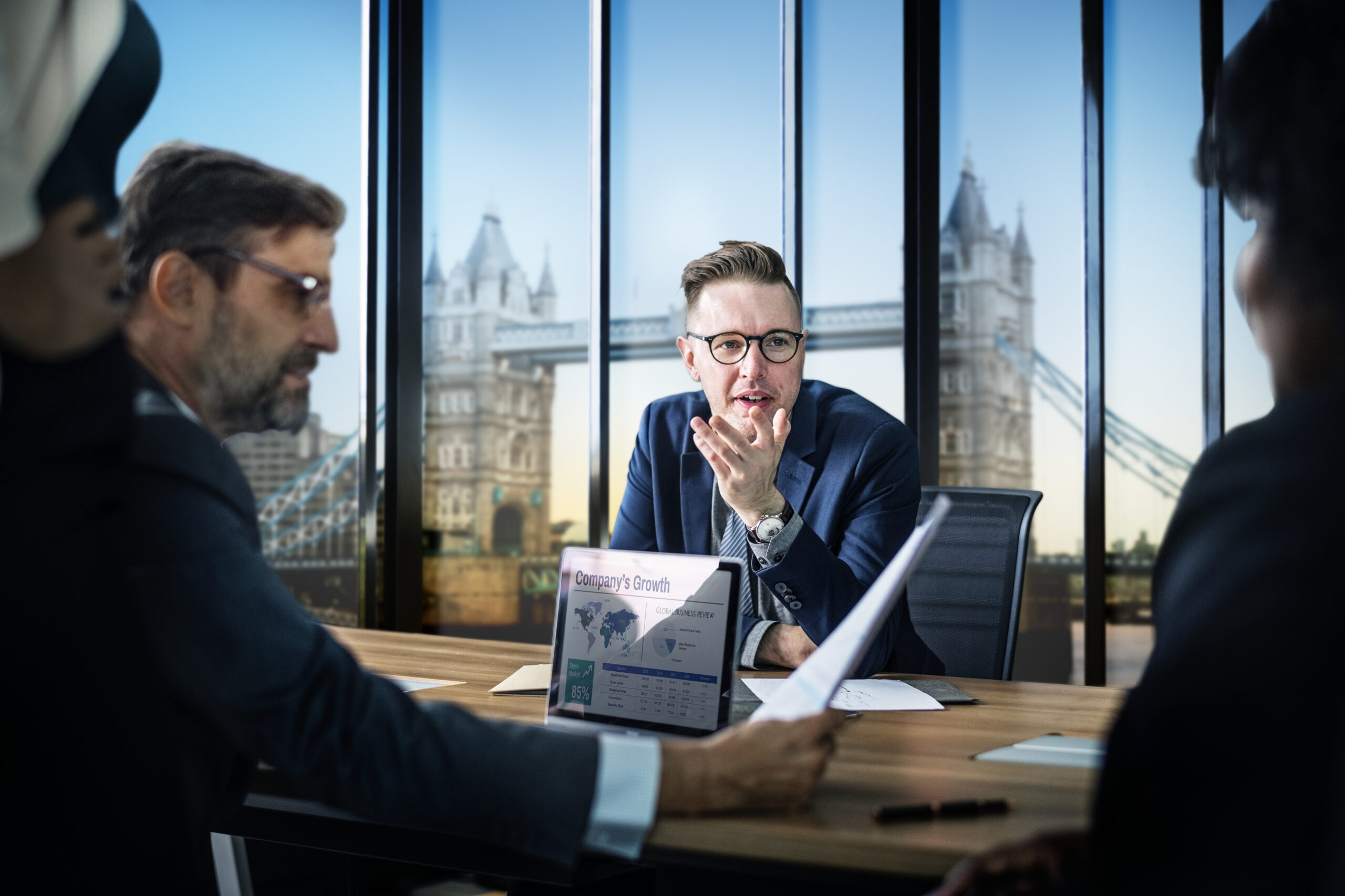 "Business executives discussing corporate strategy in a modern London office with Tower Bridge view, reviewing company growth reports during a professional boardroom meeting."