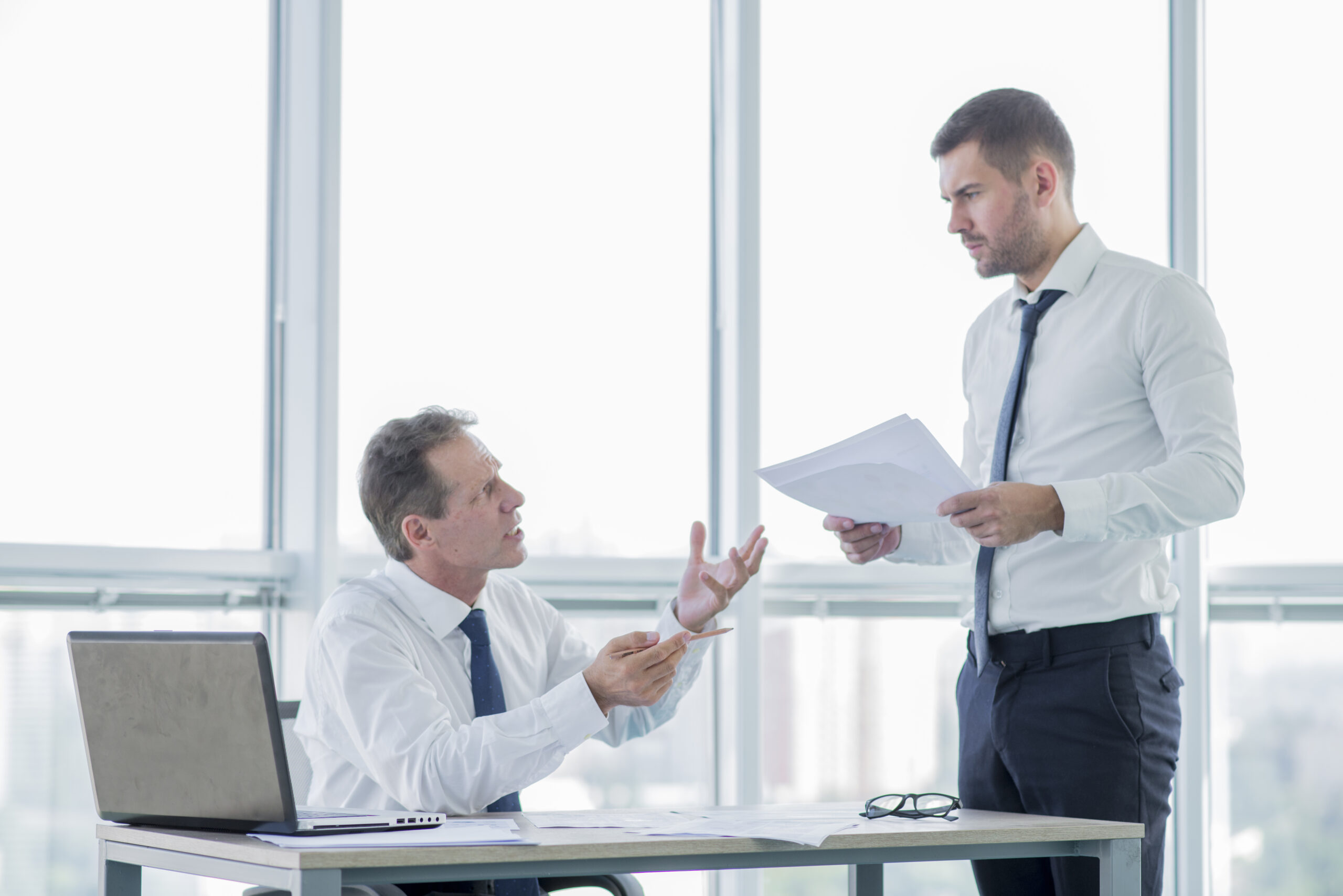 Businessmen reviewing disputed paperwork during a breach of contract disagreement in a high-rise office setting.