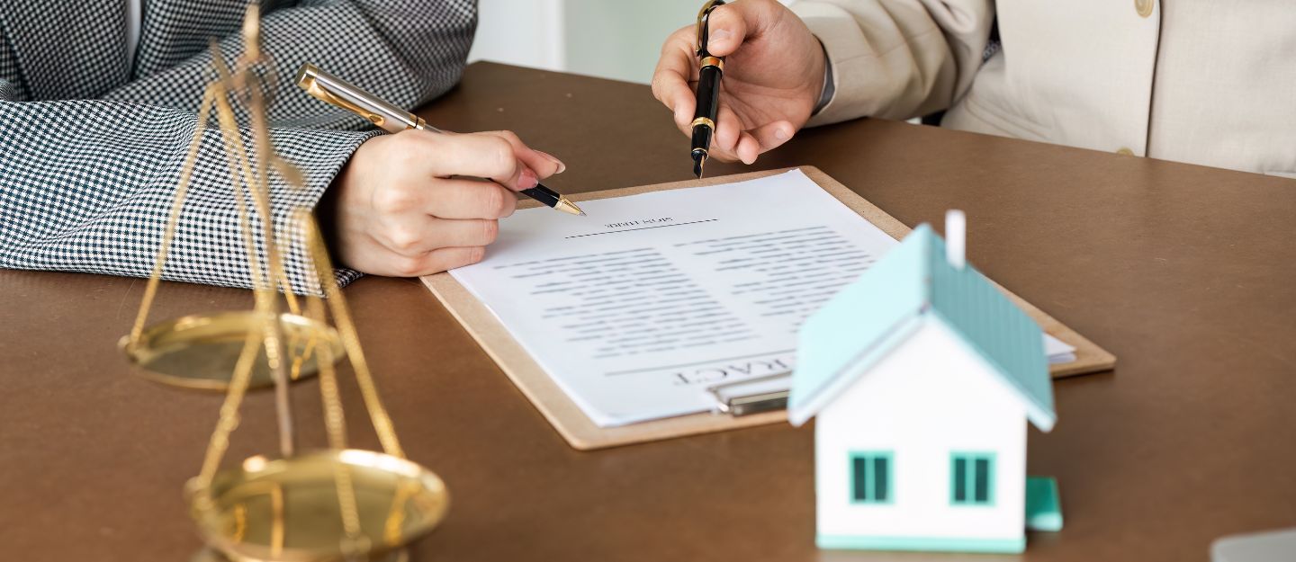 Clients signing a real estate purchase agreement during a property closing with legal documents, a house model, and scales of justice on the desk.