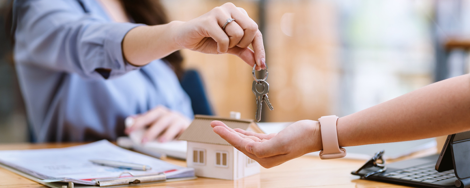 Real estate agent handing over house keys to a new homeowner during a property purchase closing with a model home and contract documents.