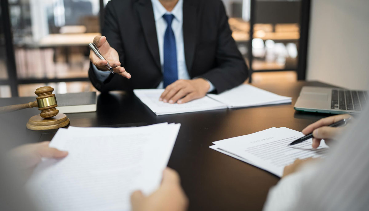A professional consultation scene showing an attorney extending a pen toward a client's contract on a desk, with a judge's gavel and laptop in the background, representing legal services and contract signing.